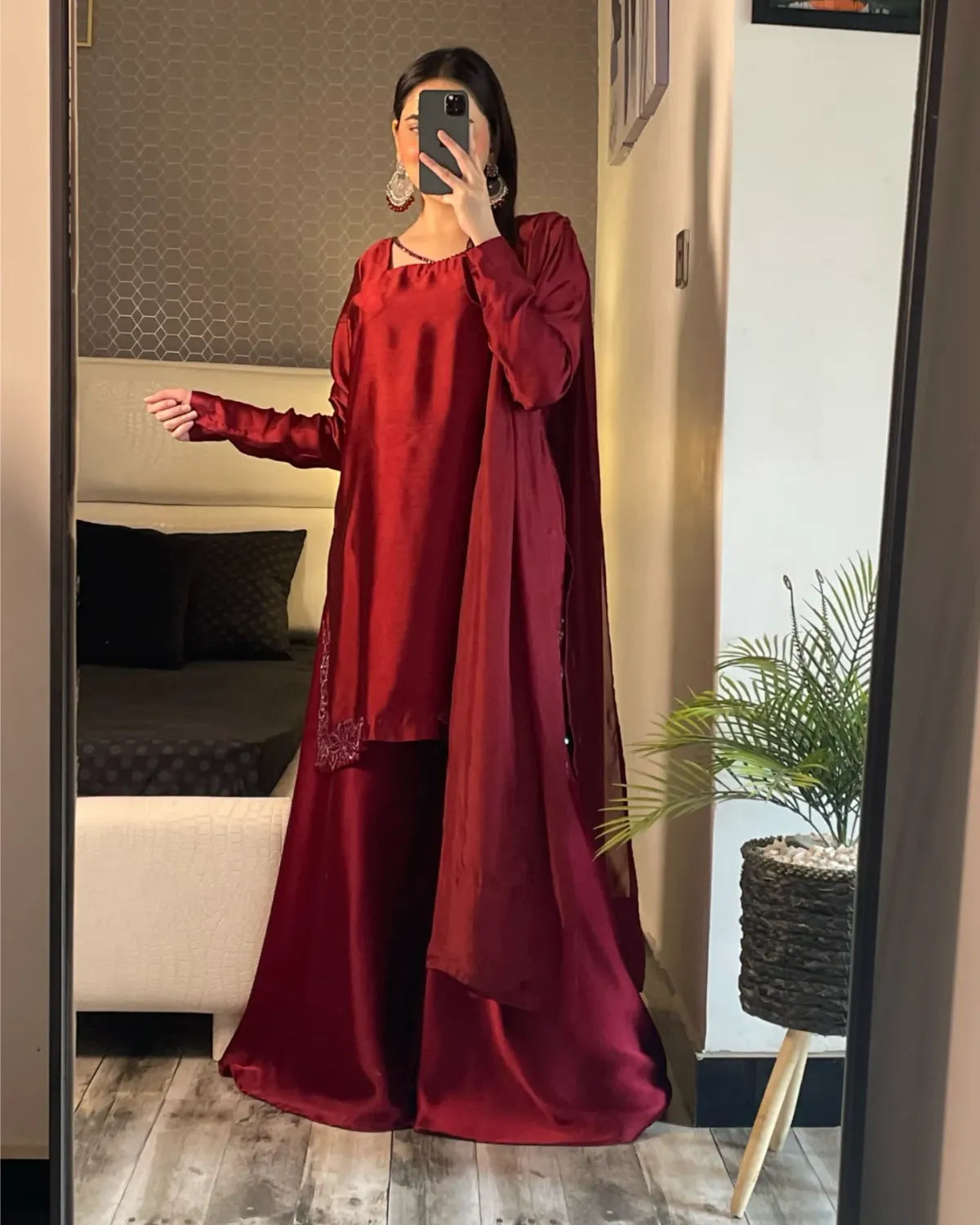 A woman takes a mirror selfie in a bedroom showing a deep red satin traditional outfit with a matching draped scarf. The outfit features a long-sleeved kurta with subtle embroidery on the hem, matching wide-leg trousers, and a draped scarf.