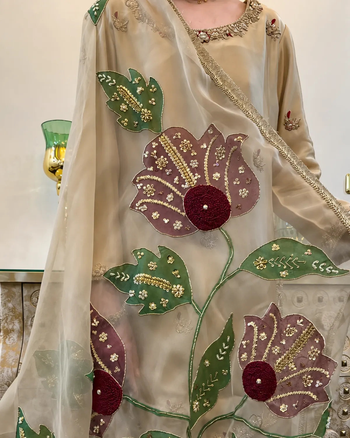 A close-up of a beige embroidered traditional garment with a sheer dupatta, featuring large floral motifs in green and mauve with gold and red embroidery.