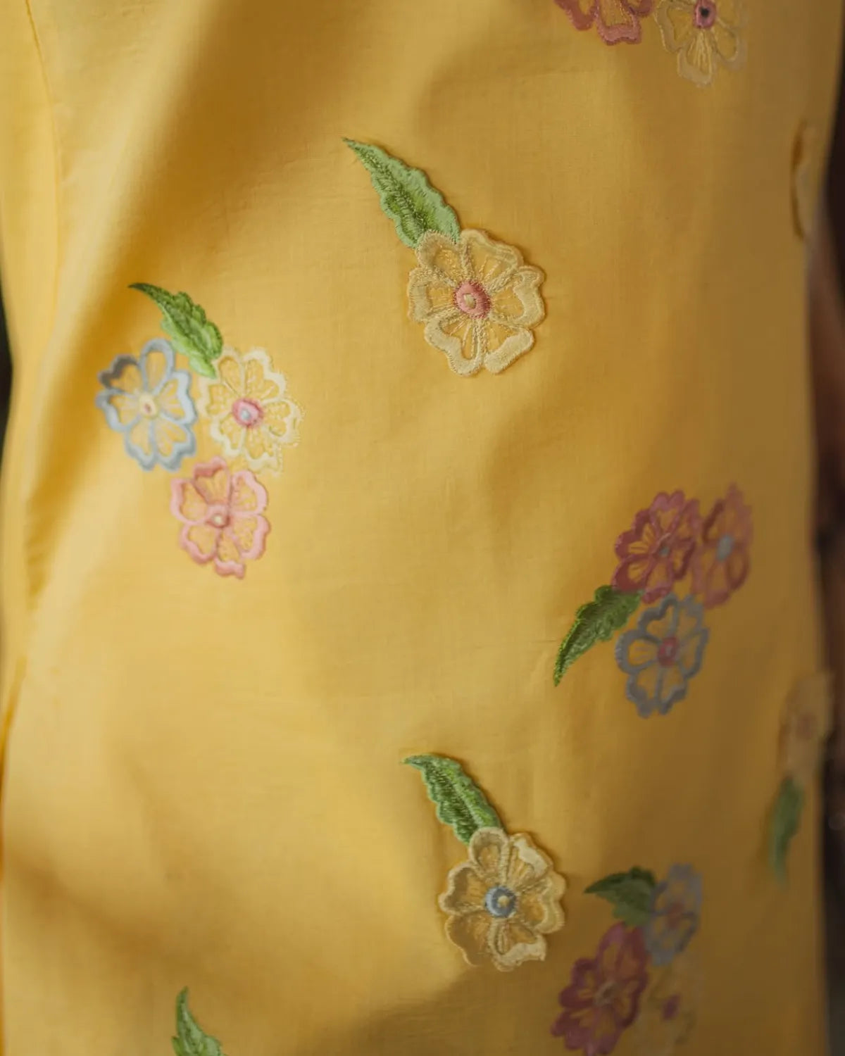 A close-up of a yellow garment featuring embroidered floral motifs in pink, blue, green, and cream.
