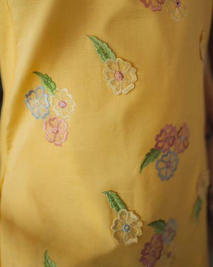 A close-up of a yellow garment featuring embroidered floral motifs in pink, blue, green, and cream.
