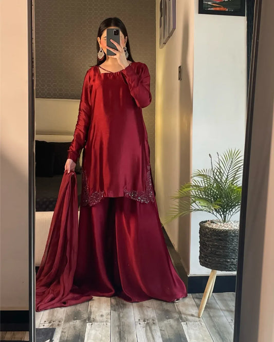 A woman takes a mirror selfie wearing a deep maroon red long-sleeve kurta and matching wide-leg trousers with a matching dupatta. The outfit features subtle shimmering embroidery near the hem.