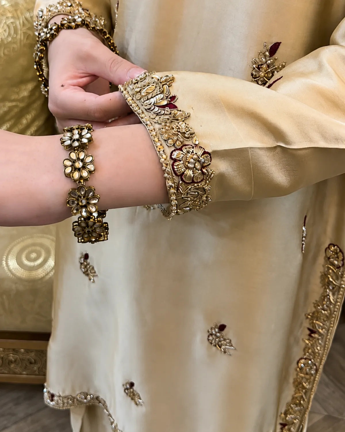 Close-up of a person wearing an ornate cream-colored embroidered traditional garment with gold floral sleeve detailing and matching gold bangles on the wrist.