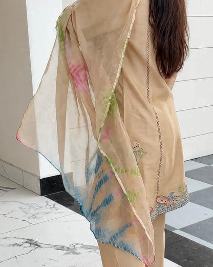A woman wearing a beige three-piece salwar kameez suit with a matching sheer dupatta featuring pink and green tie-dye patterns and blue border accents, standing in a modern indoor setting with a white paneled wall and checkered floor.
