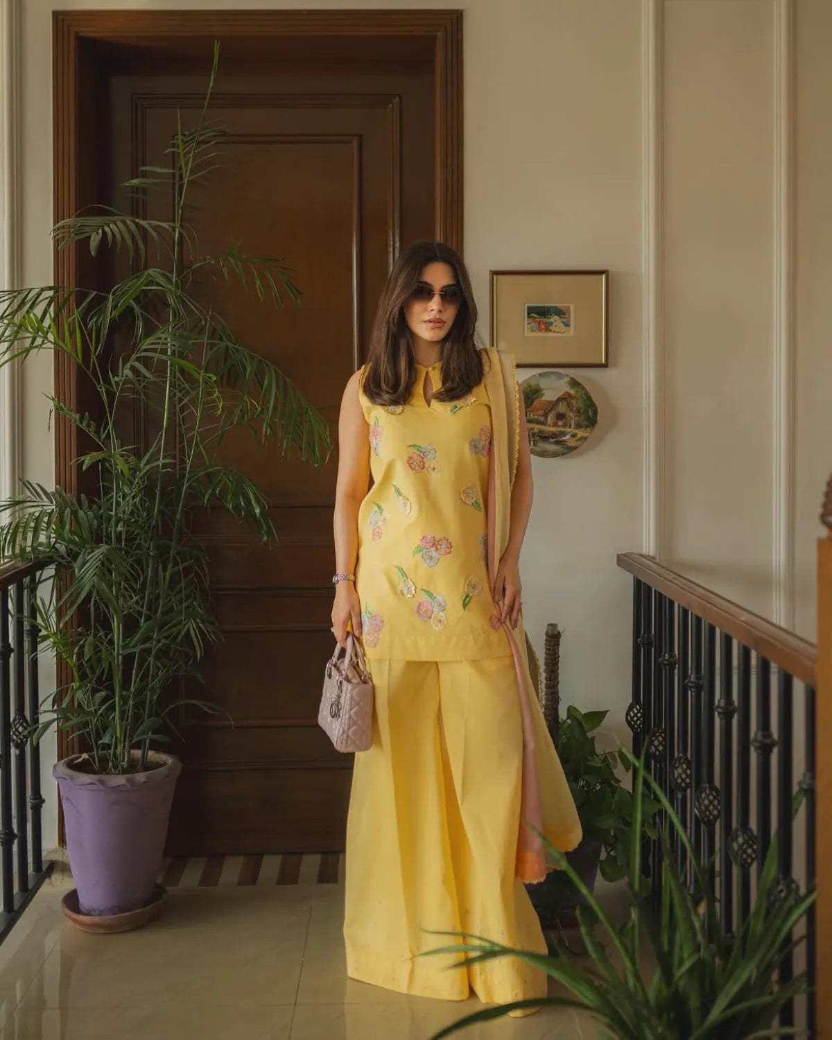 A woman stands in an indoor hallway wearing a bright yellow traditional outfit with floral embroidery, holding a small pink handbag. The setting includes a wooden door, potted plants, framed wall art, and a staircase railing.