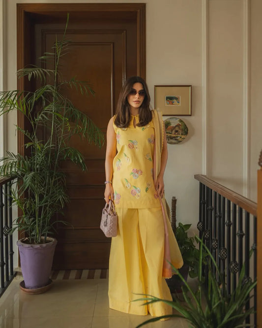 A woman stands in an indoor hallway wearing a bright yellow traditional outfit with floral embroidery, holding a small pink handbag. The setting includes a wooden door, potted plants, framed wall art, and a staircase railing.
