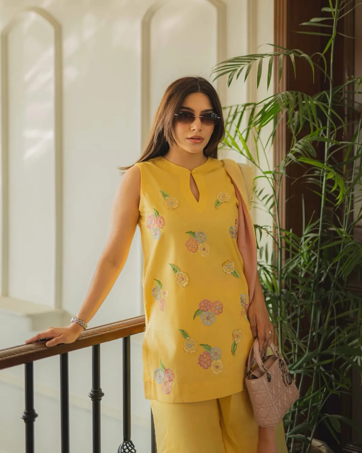 A woman wearing a yellow sleeveless kurta with colorful floral embroidery and matching yellow trousers, standing indoors near a railing and green plants.
