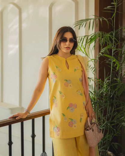 A woman wearing a yellow sleeveless kurta with colorful floral embroidery and matching yellow trousers, standing indoors near a railing and green plants.