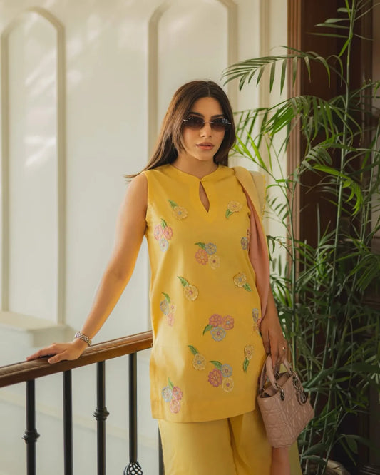 A woman wearing a yellow sleeveless kurta with colorful floral embroidery and matching yellow trousers, standing indoors near a railing and green plants.
