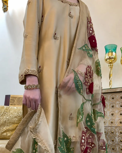 A close-up of a person wearing a beige traditional outfit with embroidered details and a matching sheer dupatta featuring red and green floral motifs with gold accents, set against an indoor background with decorative items.