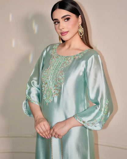 A woman modeling a mint green satin kaftan with intricate gold and green embroidery on the neckline and sleeve cuffs, set against a neutral beige background.