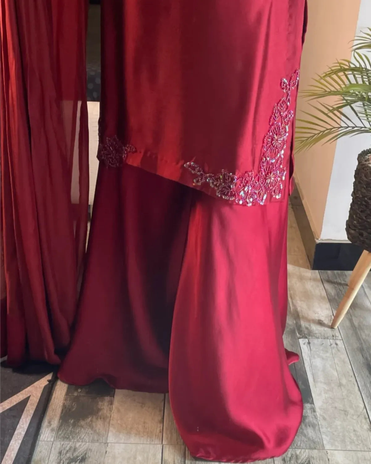 A deep red satin dress or curtain panel with silver and white beaded embroidery along the hem, shown hanging in an indoor setting with a tiled floor and a plant visible.