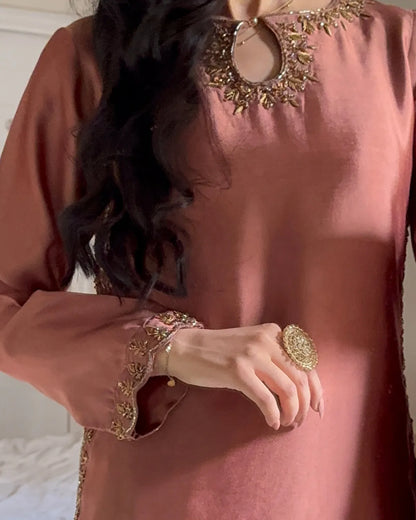 A close-up of a woman wearing a dusty rose long-sleeve tunic with ornate gold embroidery around the neckline and cuffs, paired with a large gold ring.