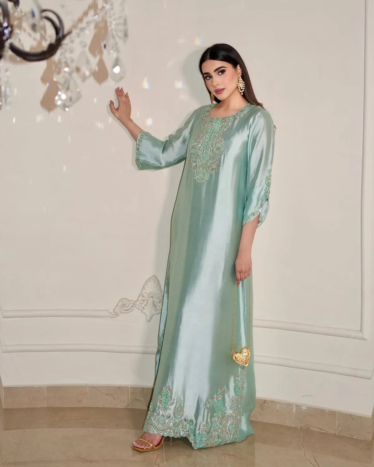 A woman models a floor-length mint green satin kaftan with gold embroidery, standing in an indoor setting with a cream-colored wall and tiled floor.