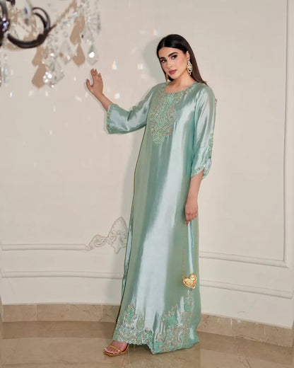 A woman models a floor-length mint green satin kaftan with gold embroidery, standing in an indoor setting with a cream-colored wall and tiled floor.