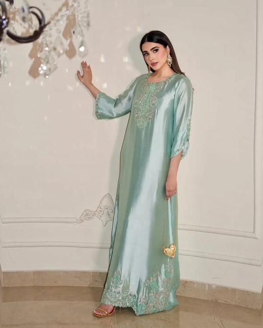 A woman models a floor-length mint green satin kaftan with gold embroidery, standing in an indoor setting with a cream-colored wall and tiled floor.