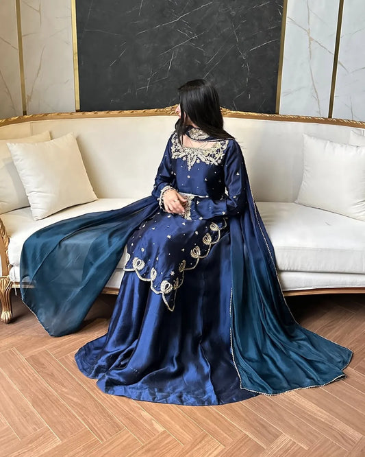 A woman wearing a deep blue traditional gown with gold embroidery sits on a cream-colored sofa in a modern living room setting.
