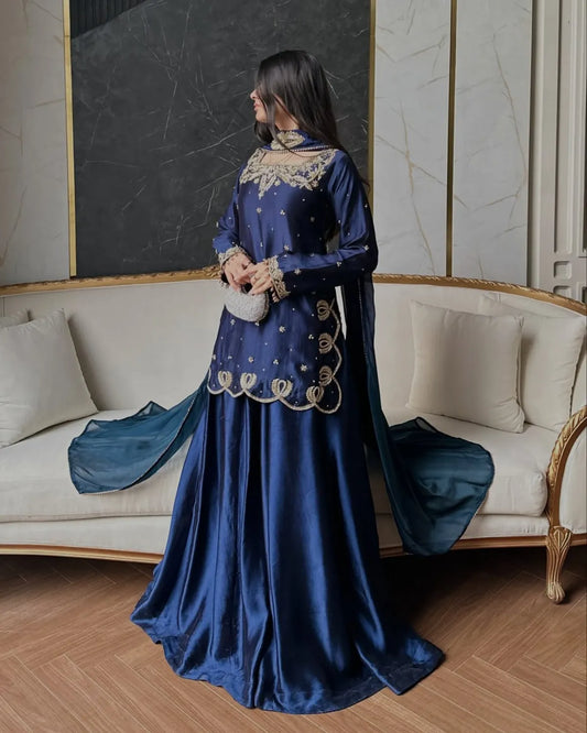 A woman wearing a long royal blue satin traditional dress with gold embroidery, standing in a luxurious room with a cream-colored sofa and marble walls.