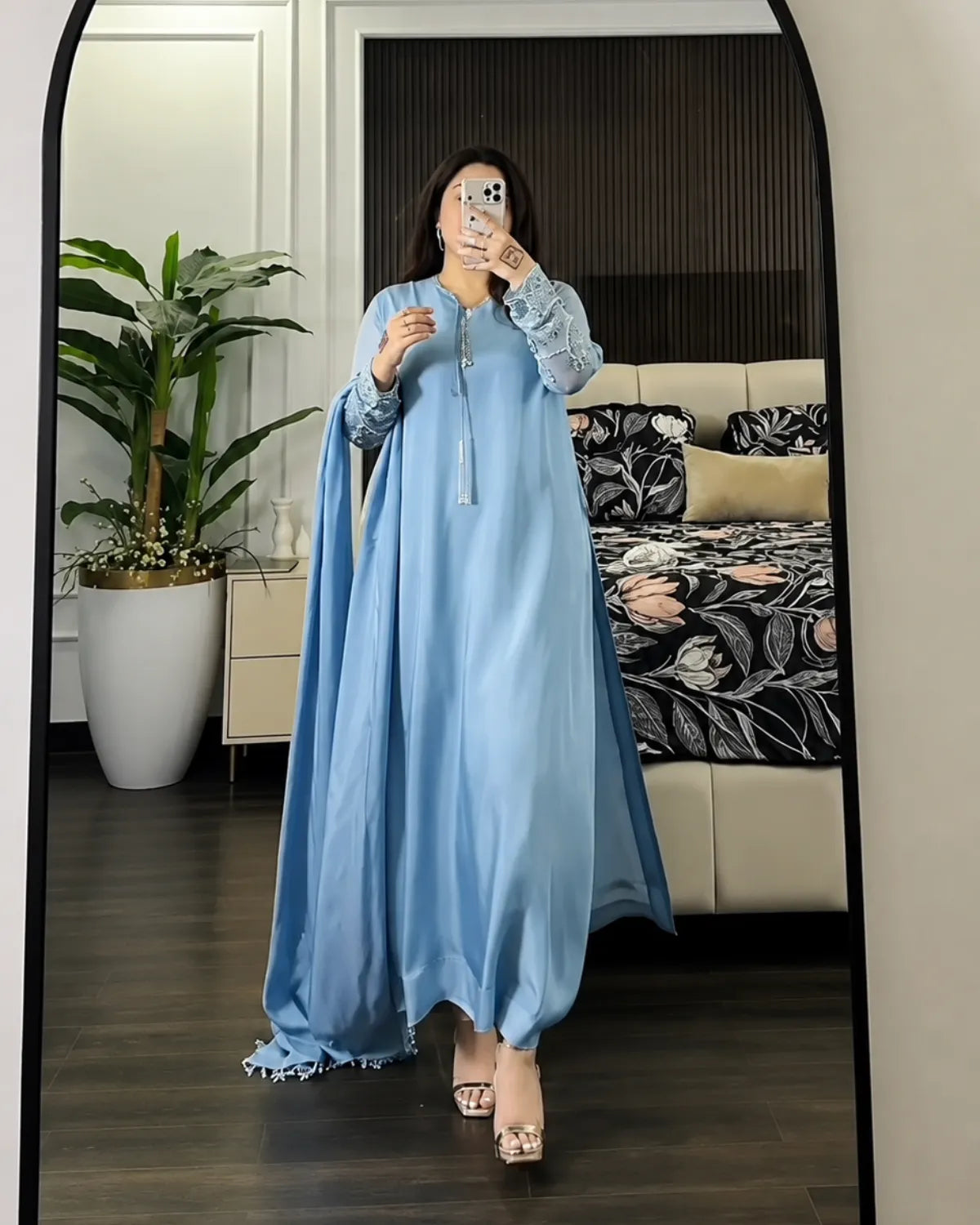 A woman takes a mirror selfie in a bedroom wearing a light blue kaftan dress with lace sleeves and a matching headscarf. The room features a bed with a black and white floral pattern, a potted plant, and dark wood flooring.