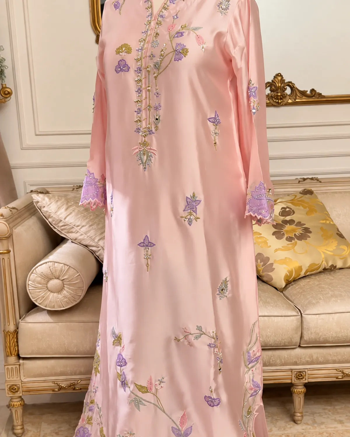 A light pink traditional long dress with purple and green floral embroidery, shown on a mannequin in an elegant cream-colored room with a sofa in the background.