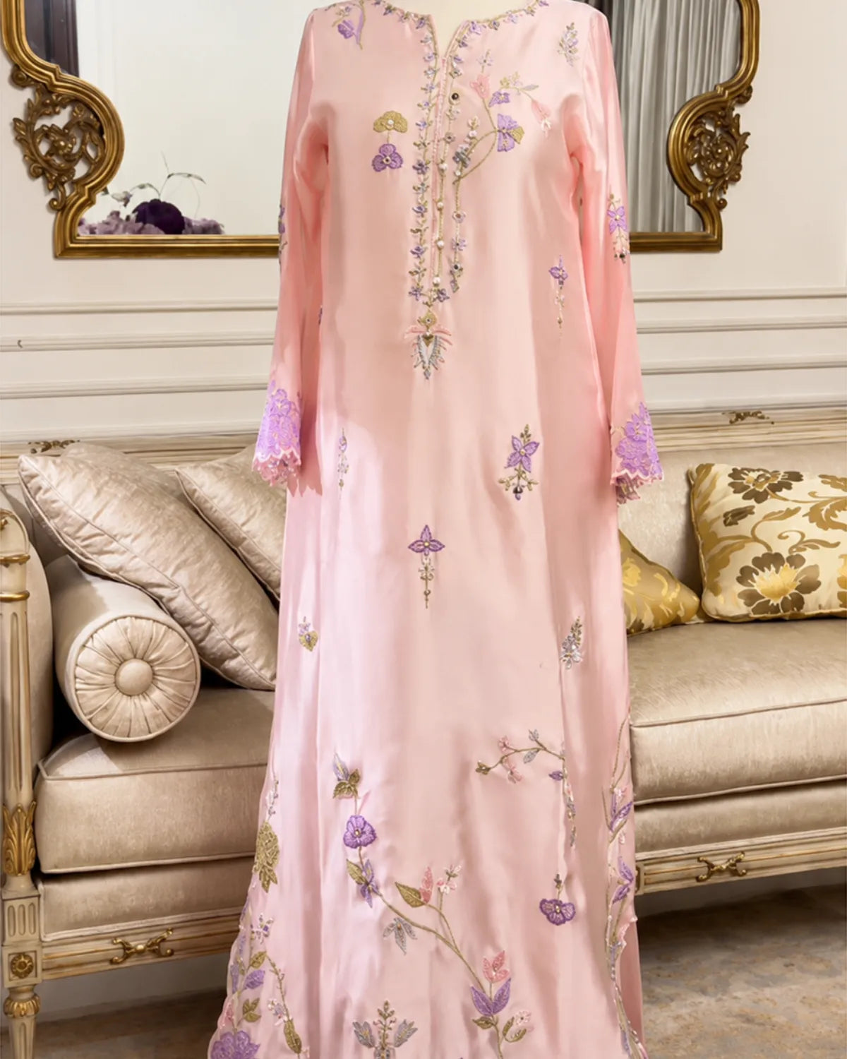 A long pink embroidered dress or caftan with floral and geometric motifs in purple, green, and gold, displayed on a mannequin in front of an ornate beige sofa and mirror.