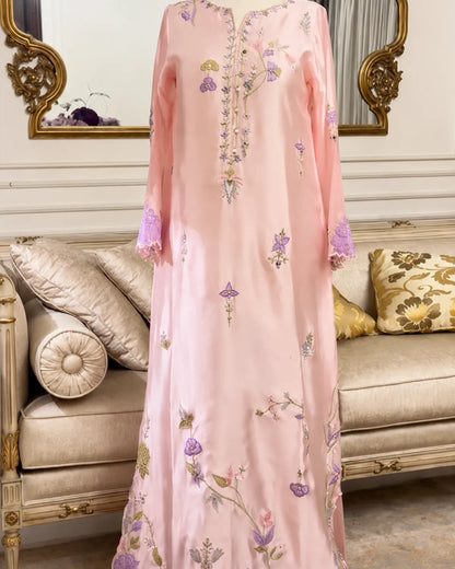 A long pink embroidered dress or caftan with floral and geometric motifs in purple, green, and gold, displayed on a mannequin in front of an ornate beige sofa and mirror.