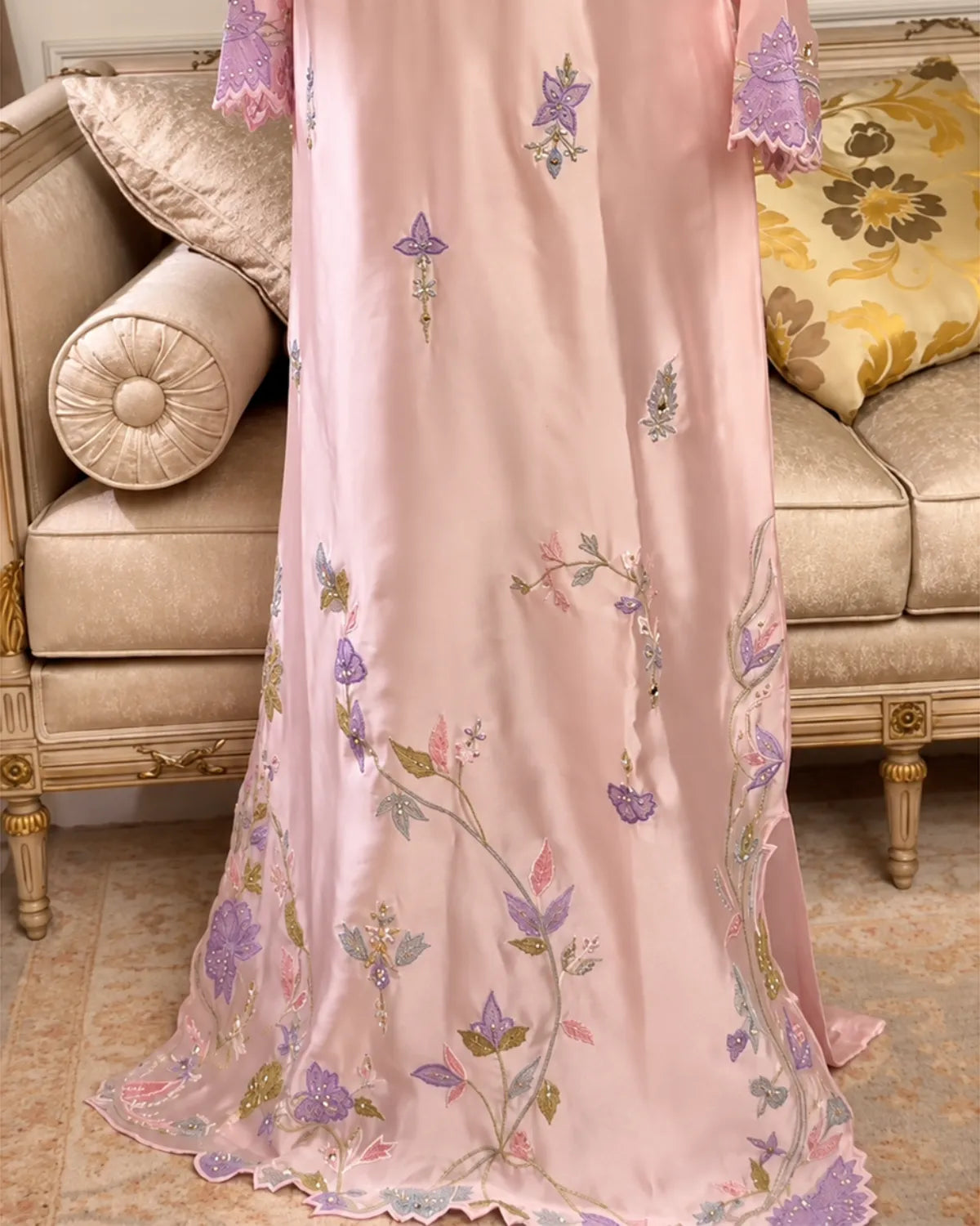 A light pink embroidered gown or dress with purple, green, and gold floral and butterfly motifs, draped over a beige sofa in an elegant indoor setting.
