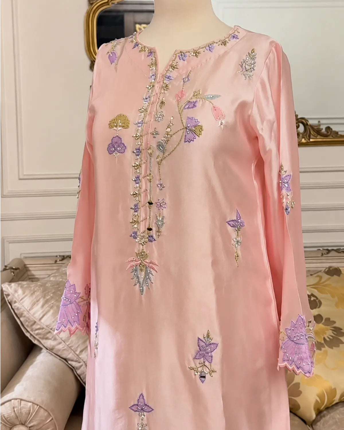 A light pink embroidered tunic dress displayed on a mannequin in an indoor setting. The tunic features purple and gold floral embroidery with scalloped cuffs.