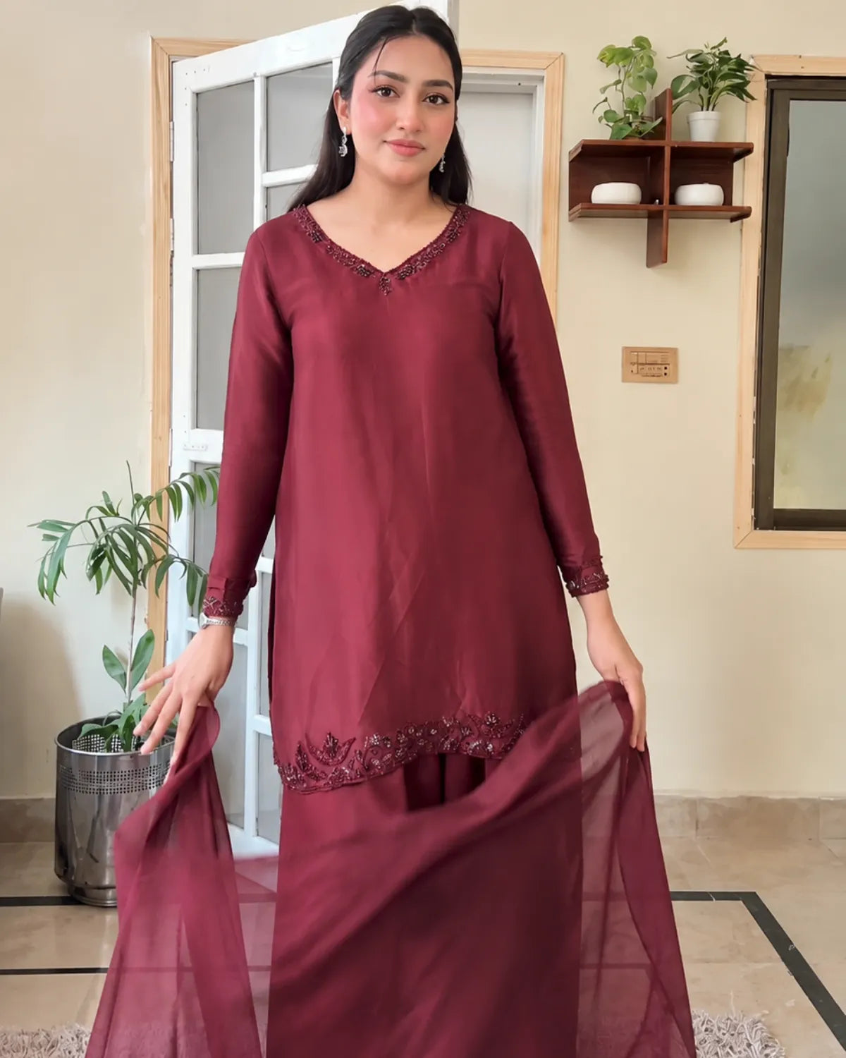 A woman modeling a three-piece maroon salwar kameez set with a matching sheer dupatta, standing in a home interior setting.