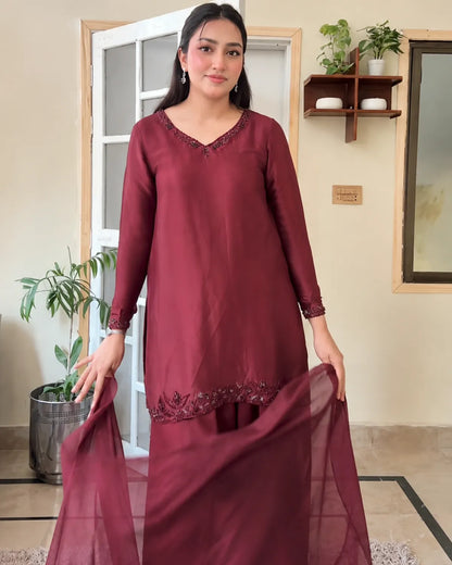 A woman modeling a three-piece maroon salwar kameez set with a matching sheer dupatta, standing in a home interior setting.