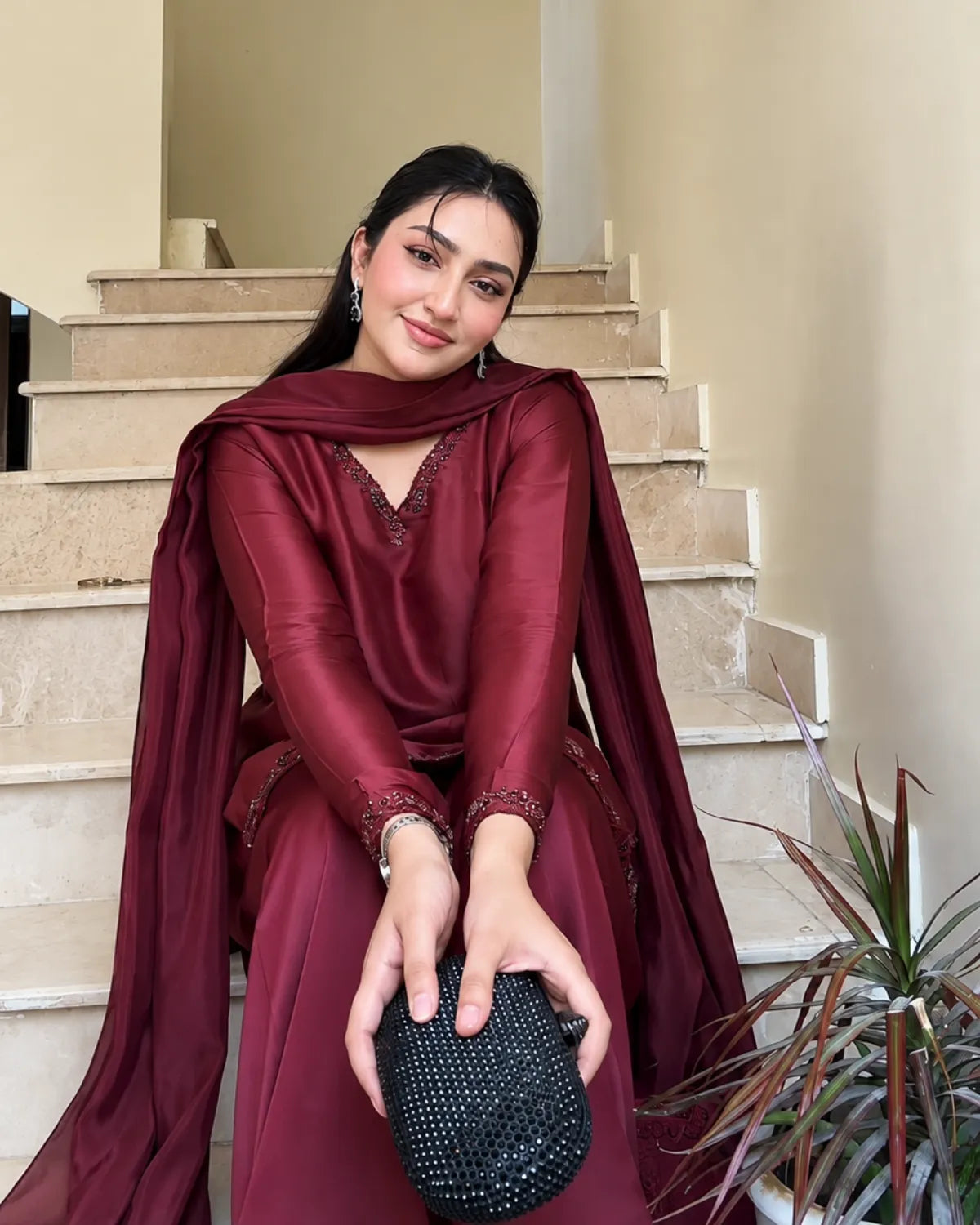 A woman wearing a deep maroon satin shalwar kameez suit with a matching dupatta, sitting on beige stone stairs and holding a black beaded clutch.