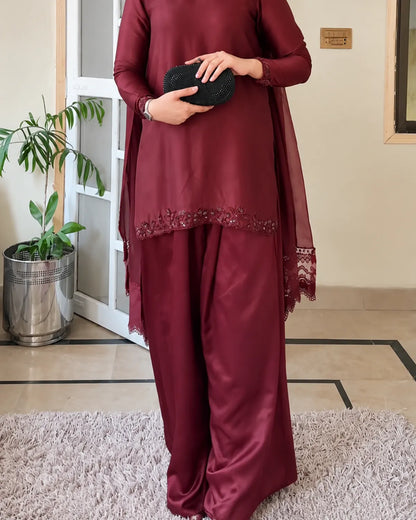 A person wearing a deep maroon three-piece traditional outfit with matching pants, a sheer dupatta, and embroidered detailing near the hem, holding a black clutch in an indoor setting.