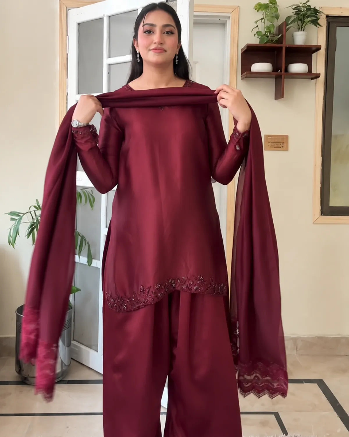 A woman modeling a deep maroon three-piece traditional outfit with a matching dupatta, standing in an indoor setting with a beige wall and wooden door in the background.