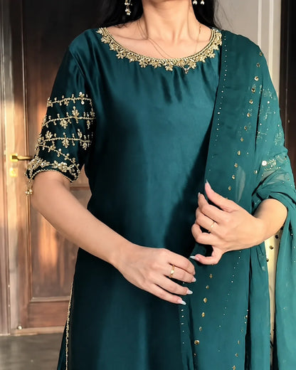 A woman wearing a dark teal green ethnic kurta with gold embroidery on the neckline and sleeves, paired with a matching sheer dupatta decorated with gold mirror work.