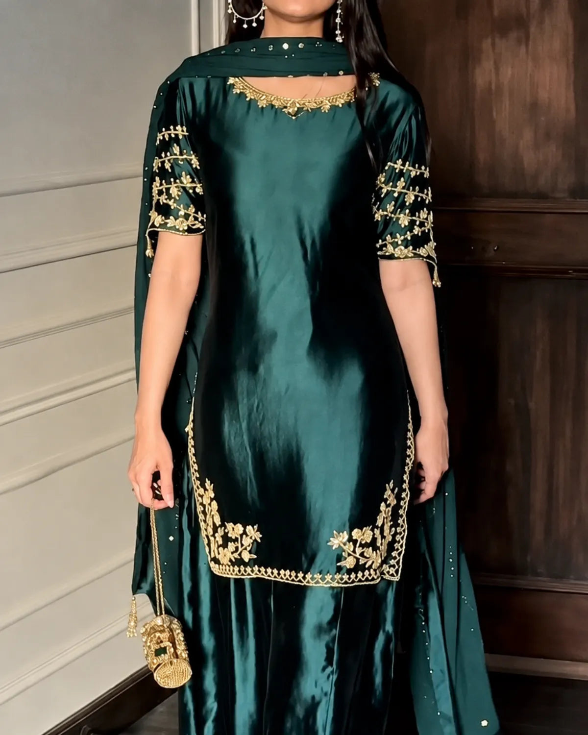 A woman wearing a dark teal green satin kurta with gold embroidery on the neckline, sleeve cuffs, and hem, paired with a matching sheer dupatta.