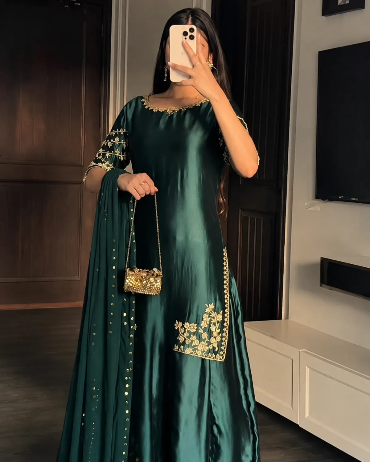 A woman takes a mirror selfie wearing a dark green satin Anarkali gown with gold floral embroidery on the sleeves and hem, holding a matching green dupatta and a small gold handbag in a modern room.