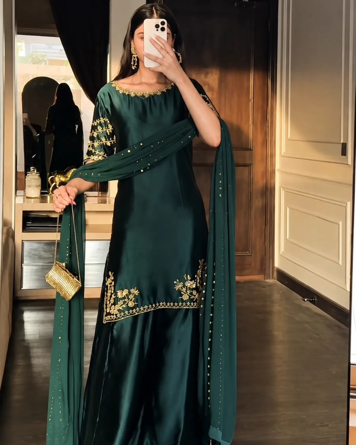 A woman takes a mirror selfie wearing a dark green traditional outfit with gold embroidery on the neckline, sleeves, and hem, paired with a matching dupatta and holding a small gold handbag.