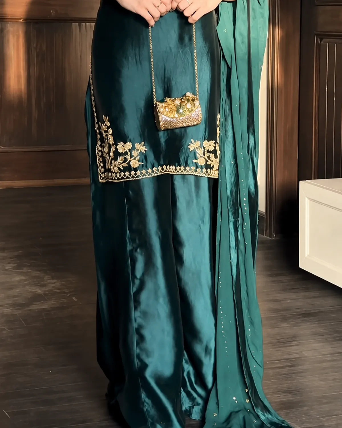 A person wearing a dark teal satin traditional outfit with gold floral embroidery near the hem, holding a small gold embellished handbag in an indoor setting with dark wood paneling.
