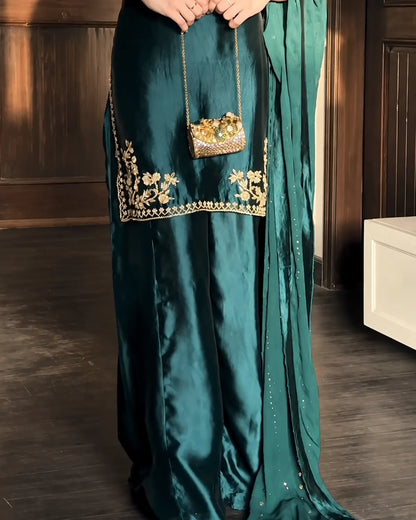 A person wearing a dark teal satin traditional outfit with gold floral embroidery near the hem, holding a small gold embellished handbag in an indoor setting with dark wood paneling.