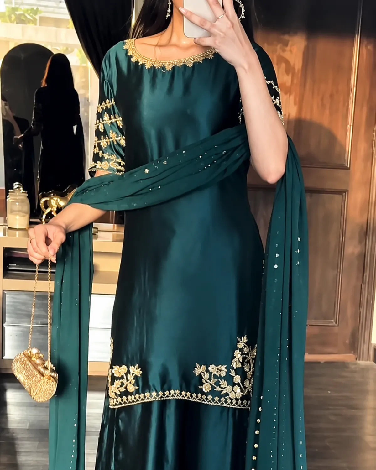 A woman wearing a dark teal green traditional outfit with gold floral embroidery and a matching dupatta, holding a small gold clutch in an indoor setting.