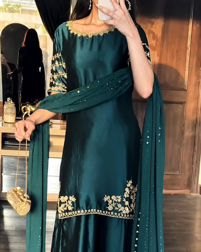 A woman wearing a dark teal green traditional outfit with gold floral embroidery and a matching dupatta, holding a small gold clutch in an indoor setting.