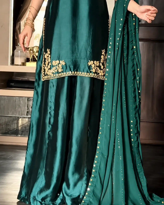 A close-up of a dark teal green traditional outfit with gold floral embroidery on the border of the tunic and a matching embroidered dupatta, shown on a model indoors.