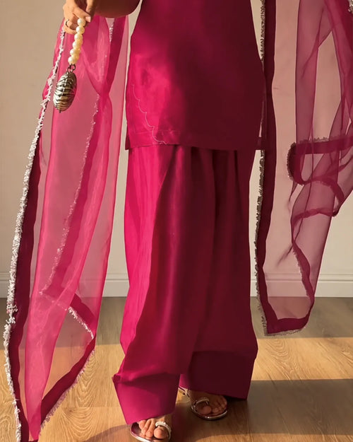 A person wearing a bright magenta pink traditional outfit with matching sheer dupatta and wide-leg pants, standing on a wooden floor.