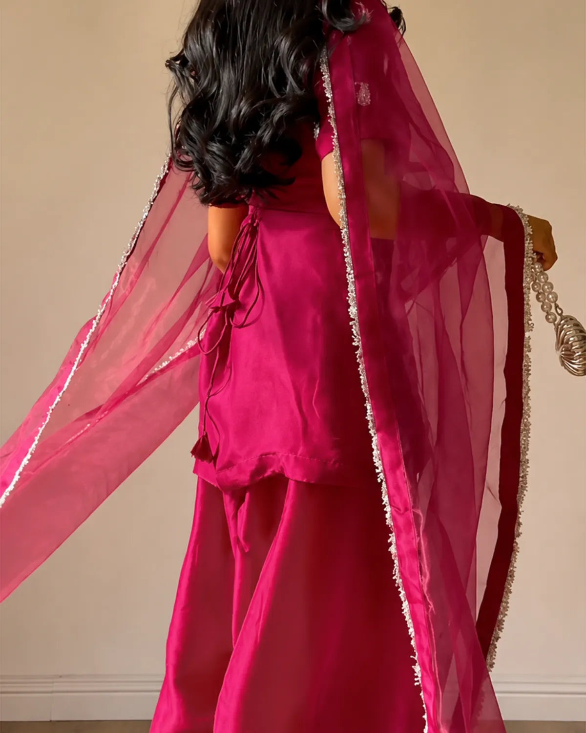 A woman shown from behind wearing a bright fuchsia pink traditional outfit with a matching sheer dupatta and a gold beaded accessory, set against a plain beige wall.