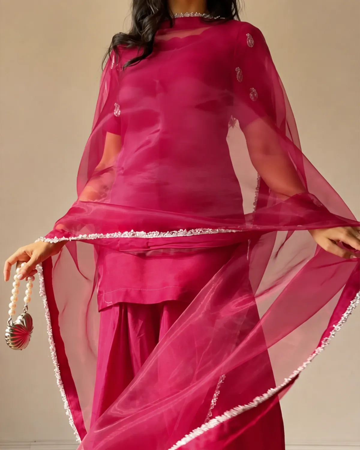 A woman wearing a bright magenta pink traditional outfit with a matching sheer organza dupatta draped over her shoulders, holding it out to show the fabric and silver embroidered border. She is holding a small silver shell purse with a beaded strap.
