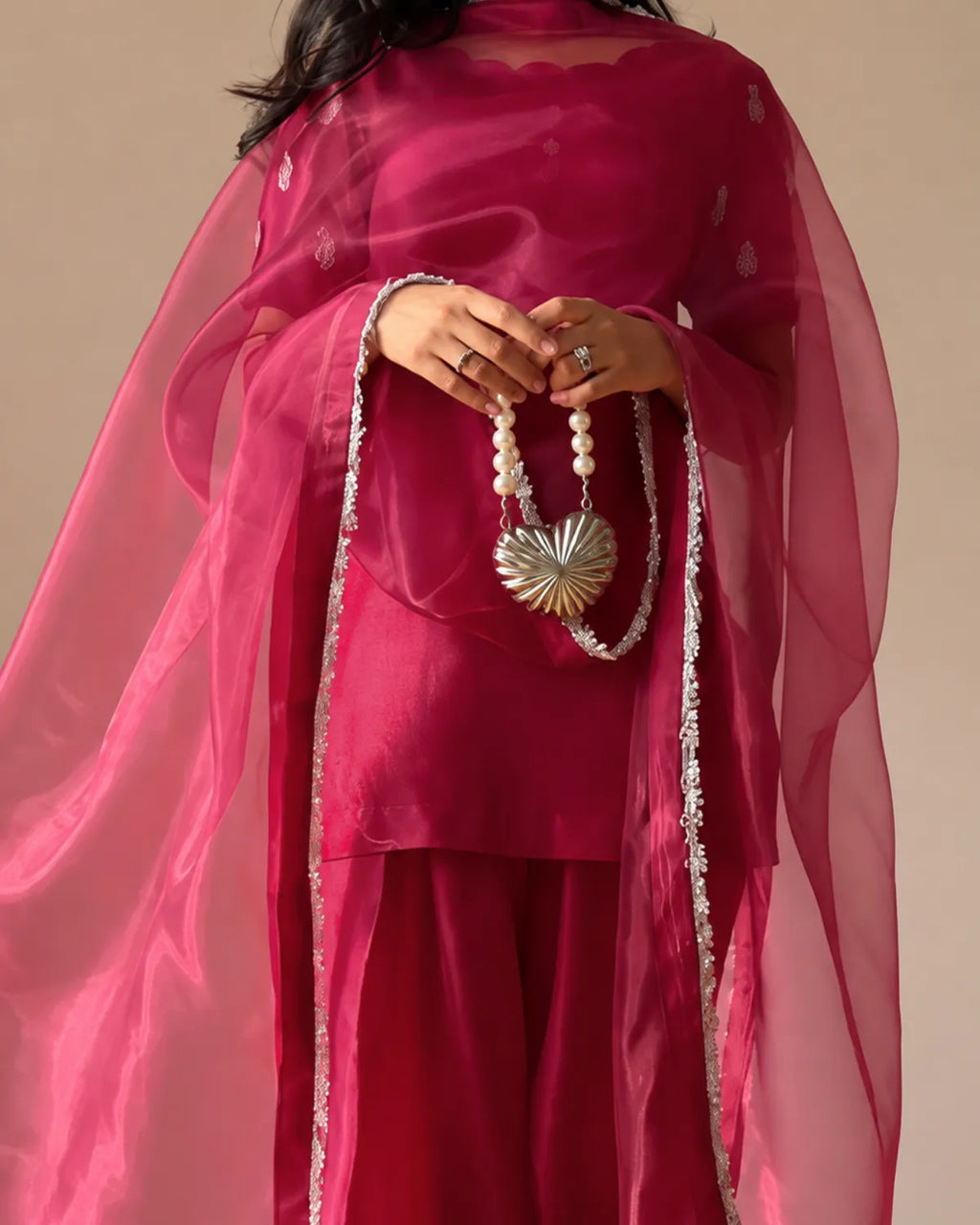 A woman wearing a bright magenta pink traditional outfit with a sheer matching dupatta and silver embroidered borders, holding a pearl beaded necklace with a metallic sun-shaped pendant against a plain beige background.