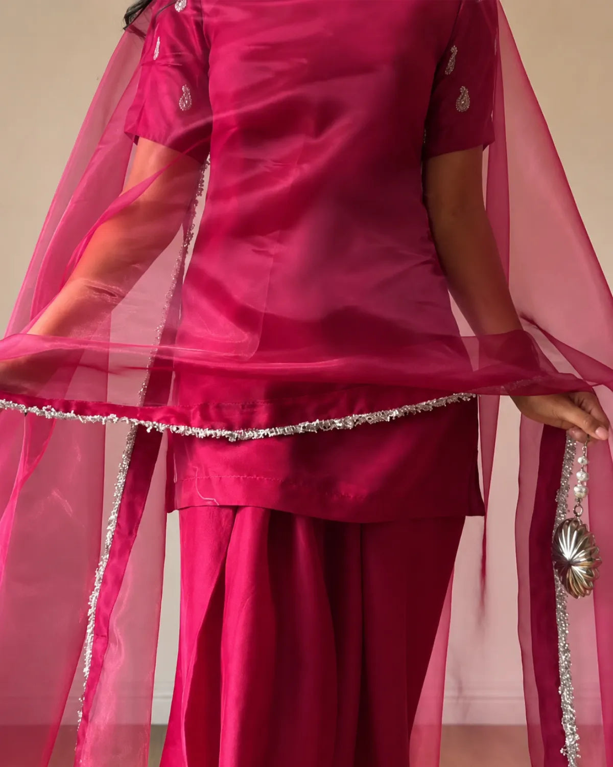 A close-up of a person wearing a bright magenta pink traditional outfit with a matching sheer dupatta. The outfit features silver embroidered details on the sleeve cuffs and a decorative silver border along the hem of the top, paired with matching pink pants.
