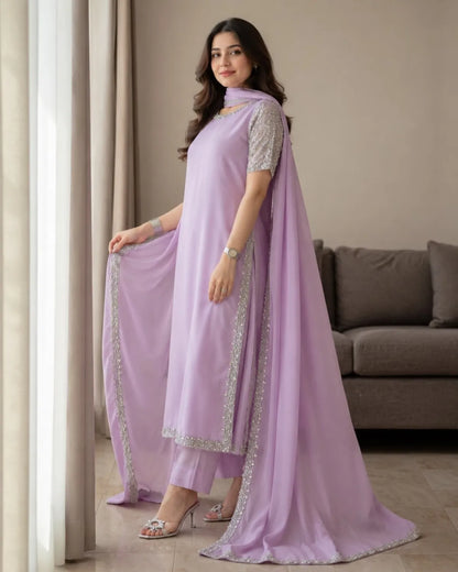 A woman models a lavender purple ethnic salwar kameez suit with matching pants and a long draped dupatta, featuring silver mirror work embroidery along the borders and sleeve edges, set in a living room environment.