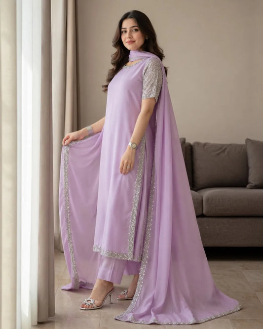 A woman models a lavender purple ethnic salwar kameez suit with matching pants and a long draped dupatta, featuring silver mirror work embroidery along the borders and sleeve edges, set in a living room environment.