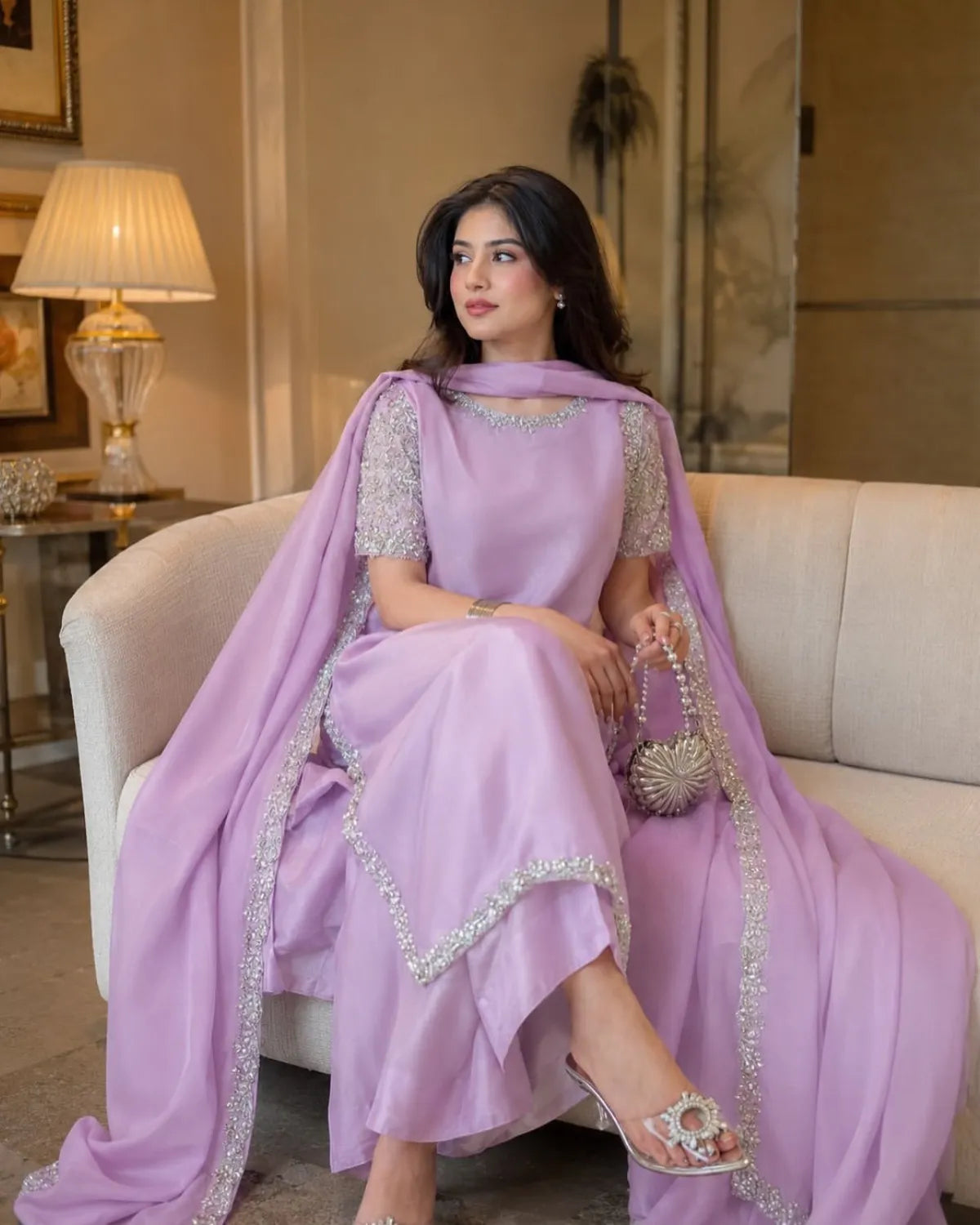 A woman seated on a cream sofa wearing a lavender traditional outfit with silver embellished borders and a matching draped scarf. She holds a small metallic handbag in a warmly lit indoor setting.
