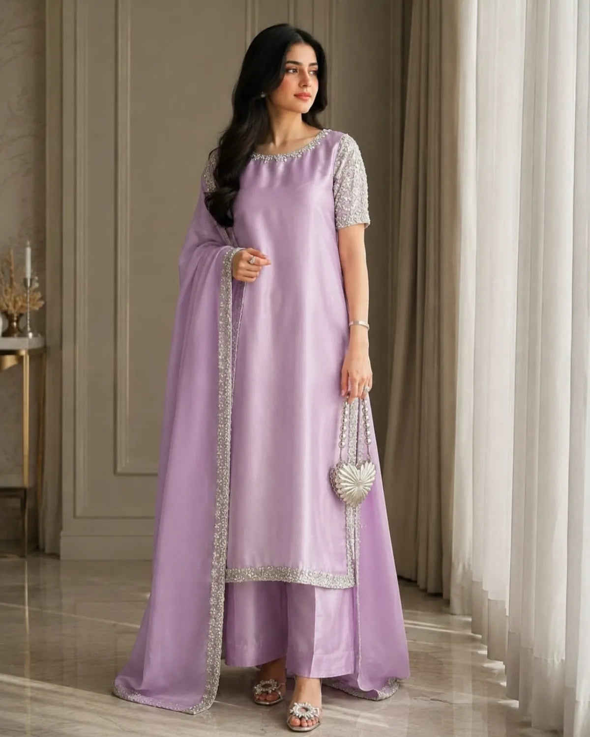 A woman models a lavender purple traditional Indian outfit, featuring a kurta with silver mirror work embroidery on the sleeves and hem, matching trousers, and a draped dupatta. She is standing in a room with beige curtains and a marble floor.